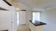 This single-story residence features an open-concept layout that connects the kitchen and living areas, creating a cohesive and functional flow throughout the space. The kitchen is equipped with white cabinetry, including a tall pantry-style cabinet that provides ample storage. A center island with a dark countertop serves as a practical workspace and visual focal point, contrasting neatly against the white lower cabinets. A white refrigerator is positioned along the kitchen wall, complementing the clean, bright aesthetic of the space. Crown molding accents the ceilings, adding a refined architectural detail throughout. The adjoining living area benefits from a large window dressed with vertical blinds, allowing natural light to filter into the space and creating an airy, open atmosphere. Light-toned flooring runs throughout, contributing to the overall brightness of the interior. The neutral color palette of warm whites and beiges lends the space a clean and welcoming ambiance. A doorway visible from the kitchen offers access to additional areas of the residence, reinforcing the practical and well-connected layout of the floor plan.