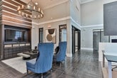 Located at 950 Battery Ave SE in Atlanta, Georgia, this single-floor amenity and coworking space offers a sophisticated and functional environment designed to support productivity and collaboration. The main open area features soaring ceilings, dark herringbone-patterned tile flooring, and a generous layout that accommodates both focused work and casual interaction. Vibrant blue velvet accent chairs, a statement chandelier, and a wall-mounted television anchor a stylish lounge area, while long shared work surfaces with ergonomic seating define a dedicated coworking zone. Decorative artwork and motivational signage contribute to an energetic, yet refined atmosphere. Multiple private offices and meeting rooms branch off from the main area, each appointed with carpeted floors, wall-mounted screens, and contemporary furnishings including wooden desks and upholstered seating. Bold accent walls in deep teal and black-and-gold artwork add visual interest to these intimate spaces. Semi-private seating nooks with high-backed upholstered chairs and side tables provide quieter spots for focused work or small conversations. Black-framed glass partition walls and doors throughout maintain visual openness while defining distinct functional zones. Natural light filters in through multiple windows, offering views of the surrounding Atlanta skyline and enhancing the overall welcoming ambiance of the space.