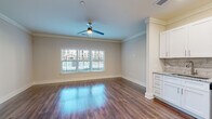 This single-floor residence combines modern finishes with a functional, open layout. Rich hardwood-style flooring flows throughout the main living areas, reflecting natural light that streams in through a large triple-pane window with exterior views. Crown molding and a ceiling fan add refined detail to the spacious living area, which connects seamlessly to the kitchen.

The kitchen is well-appointed with crisp white shaker-style cabinetry, granite countertops, and a marble-patterned tile backsplash. A full suite of stainless steel appliances includes a French door refrigerator with ice maker, electric range, over-the-range microwave, and dishwasher. A decorative diamond-patterned tile accent and upper glass-front cabinetry add visual interest.

The bathroom features hardwood-style flooring consistent with the rest of the unit, a soaking tub with a subway tile surround extending to the ceiling, brushed nickel fixtures, and a curved shower rod. The neutral, warm-toned walls throughout the unit create a cohesive, welcoming ambiance well-suited to a variety of interior styles.