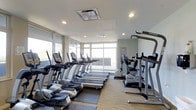 This fitness center offers a bright and modern exercise environment designed to support a full cardio workout routine. Large floor-to-ceiling windows wrap around the space, flooding the room with abundant natural light and providing open views of the surrounding exterior — creating an energizing atmosphere for workouts. The clean, contemporary design features a neutral color palette of whites and soft grays, complemented by light flooring that adds to the airy feel of the space. The facility is outfitted with a well-curated lineup of True-brand cardio equipment, including multiple treadmills, elliptical cross-trainers, and upright stationary bikes, all arranged to maximize usability and flow throughout the room. A dedicated strength training station is also present, mounted along the mirrored wall, which further enhances the sense of openness and allows users to monitor their form. Recessed lighting in the ceiling supplements the natural daylight, ensuring the space remains well-lit at all hours. The overall ambiance is clean, professional, and welcoming — providing an inviting environment for residents or guests looking to maintain an active lifestyle without leaving the building.