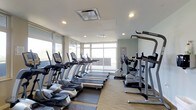 This fitness center offers a bright and modern exercise environment designed to support a full cardio workout routine. Large floor-to-ceiling windows wrap around the space, flooding the room with abundant natural light and providing open views of the surrounding exterior — creating an energizing atmosphere for workouts. The clean, contemporary design features a neutral color palette of whites and soft grays, complemented by light flooring that adds to the airy feel of the space. The facility is outfitted with a well-curated lineup of True-brand cardio equipment, including multiple treadmills, elliptical cross-trainers, and upright stationary bikes, all arranged to maximize usability and flow throughout the room. A dedicated strength training station is also present, mounted along the mirrored wall, which further enhances the sense of openness and allows users to monitor their form. Recessed lighting in the ceiling supplements the natural daylight, ensuring the space remains well-lit at all hours. The overall ambiance is clean, professional, and welcoming — providing an inviting environment for residents or guests looking to maintain an active lifestyle without leaving the building.
