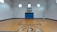 Basketball 360 is a well-appointed indoor basketball facility designed to deliver a complete court experience in a clean, functional environment. The court itself is the centerpiece of the space, featuring a polished hardwood floor finished with crisp blue and black court markings, including a prominent center-court logo. Regulation-height basketball hoops are mounted on both ends of the court, each equipped with a clear backboard and blue safety padding along the base of the wall for player protection.

The facility's white-painted cinder block walls rise to generous heights, creating an open, airy atmosphere well-suited for full gameplay. A row of arched windows positioned near the top of the walls on multiple sides allows abundant natural light to pour in, brightening the court and reducing reliance on the overhead lighting fixtures mounted along the ceiling's exposed structural framework.

The overall layout is straightforward and purpose-driven, with clearly defined court boundaries and ample out-of-bounds space on all sides. An informational plaque displayed on one wall adds a touch of character to the space. Emergency exits are clearly marked and accessible. The combination of quality flooring, proper safety features, and bright, naturally lit surroundings makes Basketball 360 a well-equipped venue for practice, training, or competitive play.