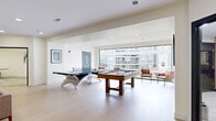 This well-appointed amenity suite spans a single floor and is thoughtfully designed to serve both recreational and professional needs. The open-plan game room is the centerpiece of the space, featuring light hardwood flooring, crisp white walls, and abundant natural light streaming in through large picture windows that offer views of the surrounding building exterior. The game room is equipped with a pool table and a ping pong table, providing ample entertainment options, while a comfortable lounge area with a sofa, accent chairs, and casual seating arrangements creates an inviting space for socializing and relaxation.

Adjacent to the game room, three private office spaces offer quiet, focused work environments. One office features an L-shaped workstation with dual computer monitors and carpeted flooring, accessed through a glass-paneled door. The remaining two offices each contain built-in desks and task seating, ideal for small meetings or individual work sessions. The offices are finished with neutral tones and patterned carpet, providing a calm and professional atmosphere.

Recessed lighting throughout the suite ensures consistent illumination across all areas, complementing the clean, modern aesthetic that defines the entire space.