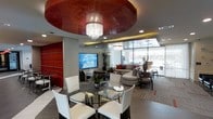 This single-floor space makes a striking first impression through its bold contemporary design and carefully curated interior elements. A dramatic circular ceiling medallion finished in deep mahogany red serves as the room's focal point, anchoring a crystal chandelier that adds an elegant, glamorous touch. A secondary chandelier echoes this detail further into the space, maintaining visual continuity throughout.

The open-plan layout seamlessly integrates a dining area and a lounge zone. The dining area features a round glass-top table surrounded by sleek white chairs, while the lounge is furnished with a generously sized sectional sofa and coordinating accent chairs in neutral tones, accented with patterned throw pillows. A large flat-screen television mounted on the wall serves as an entertainment focal point for the lounge area.

One wall showcases a textured red accent panel, adding depth and a vibrant pop of color that complements the ceiling medallion. Floor-to-ceiling windows along the perimeter flood the interior with natural light and provide clear views of the surrounding exterior. Potted tropical plants are thoughtfully placed throughout, softening the modern aesthetic and adding a refreshing organic element to the overall ambiance.