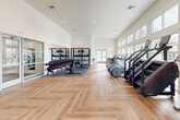 Located at 1401 Creston Rd in Paso Robles, California, this single-floor fitness center offers a generously sized, well-appointed workout facility designed to support a comprehensive exercise experience. The space is defined by soaring high ceilings and expansive floor-to-ceiling windows that line the walls, flooding the interior with abundant natural light and providing open views of the surrounding outdoor community areas.

The flooring features an elegant herringbone-patterned light wood finish, lending a clean and modern aesthetic throughout. A bold, colorful typographic fitness mural serves as a dynamic focal point on the main wall, energizing the atmosphere with motivational activity words in teal and orange tones.

The facility is equipped with a wide variety of cardio machines, including treadmills, stair climbers, and stationary bikes, arranged to take full advantage of the natural light and outdoor views. A dedicated strength training area includes cable machines, a power rack, and a well-stocked dumbbell rack with accompanying benches, all set on protective rubber flooring mats. Large wall mirrors enhance the sense of space and support proper form during workouts.

Recessed lighting supplements the natural light, ensuring the space remains bright and inviting at all hours. The overall design is modern, clean, and functional, creating an inspiring environment for fitness enthusiasts of all levels.