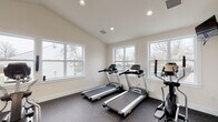 Located at 155 Southern Trace in Cincinnati, Ohio, this dedicated fitness center offers a well-equipped, single-floor workout facility designed to support a comprehensive exercise routine. The space features vaulted ceilings that lend an open, airy feel, complemented by recessed lighting throughout. Large windows line multiple walls, flooding the room with natural light and providing views of the surrounding outdoor area, including a glimpse of a nearby pool and landscaping.

The flooring consists of durable rubber gym matting, ideal for both cardio and strength training activities. The cardio section is outfitted with multiple treadmills and elliptical machines, while a stationary bike adds additional variety for low-impact workouts. A multi-station cable machine provides a range of strength training options, and a full rack of rubber hex dumbbells in graduated weights supports free weight exercises. Medicine balls and stability balls round out the functional fitness offerings.

A wall-mounted television is positioned for easy viewing during cardio sessions, and a French door entry leads to an adjacent outdoor or lobby area. The clean, neutral color palette of white and warm beige walls creates an inviting and motivating environment. Overall, this fitness center combines practical equipment, thoughtful design, and ample natural light to deliver a versatile and comfortable workout experience.