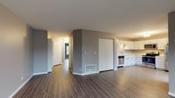 This single-floor unit is designed with a clean, contemporary aesthetic throughout. Wood-look plank flooring in warm and cool gray tones runs continuously across the open-concept living and dining area, creating a cohesive and expansive feel. The layout connects the main living space directly to the kitchen, which is outfitted with white shaker-style cabinetry, light countertops, and a full suite of stainless steel appliances including a refrigerator, electric range, built-in microwave, and dishwasher. A sliding glass door in the kitchen area provides access to an outdoor deck, allowing natural light to fill the space. A central hallway leads to the private areas of the unit, where bedrooms feature large windows that bring in additional natural light and offer views of the surrounding landscape. The walls are painted in neutral gray tones throughout, complementing the flooring and trim. A walk-in closet with wire shelving provides generous storage capacity. The full bathroom includes a single-sink vanity with white cabinetry, a framed mirror, and a tub-shower combination, finished with the same wood-look flooring for a unified design.
