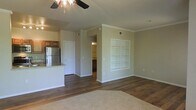 This single-floor residence offers a thoughtfully designed open-concept layout that seamlessly connects the main living area to the kitchen. The living room is finished with rich hardwood-style flooring and features a ceiling fan with a light fixture overhead, adding both comfort and functionality. A standout built-in shelving unit with a grid-style design provides both storage and a decorative focal point along one wall, while crown molding lends a refined touch throughout.

The kitchen is positioned adjacent to the living space and is equipped with stainless steel appliances, including a full-sized refrigerator, and showcases a breakfast bar with granite-style countertops and warm wood cabinetry above. The neutral, earthy wall tones create a cohesive and inviting atmosphere throughout the home.

A hallway leads from the main living area to additional rooms, with a glimpse of what appears to be a vanity area or secondary space visible in the background. The layout is efficient and practical, making excellent use of the available floor plan. Overall, the space presents a clean, move-in-ready aesthetic with durable finishes and classic design elements that appeal to a wide range of tastes.