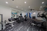 This fitness center is thoughtfully designed to support a wide range of workout routines within a single-floor layout. The space is divided into distinct functional zones, each serving a specific training purpose while maintaining a cohesive, modern aesthetic throughout.

The cardio area features multiple treadmills, elliptical machines, and stationary cycling bikes, all positioned near large windows that invite natural light and offer views of the surrounding outdoor greenery. Wall-mounted flat-screen televisions are positioned throughout for entertainment during workouts. Ceiling fans provide additional airflow and comfort across the space.

A strength and resistance training section houses a cable machine, leg press equipment, and a variety of resistance bands mounted on the wall, providing versatile options for muscle conditioning. A well-stocked dumbbell rack with a range of weights lines one wall, accompanied by medicine balls and stability balls for functional fitness exercises.

Large framed mirrors are strategically installed to visually expand the space and allow users to monitor their form. A vibrant watercolor-style mural adds an artistic focal point, lending energy and personality to the environment.

The flooring transitions between bold, patterned athletic carpet in the main training areas and smooth hard flooring in the open zones, clearly delineating workout spaces. Recessed lighting throughout ensures the entire facility is well-lit, creating a clean, professional atmosphere conducive to focused training.