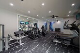 This fitness center is thoughtfully designed to support a wide range of workout routines within a single-floor layout. The space is divided into distinct functional zones, each serving a specific training purpose while maintaining a cohesive, modern aesthetic throughout.

The cardio area features multiple treadmills, elliptical machines, and stationary cycling bikes, all positioned near large windows that invite natural light and offer views of the surrounding outdoor greenery. Wall-mounted flat-screen televisions are positioned throughout for entertainment during workouts. Ceiling fans provide additional airflow and comfort across the space.

A strength and resistance training section houses a cable machine, leg press equipment, and a variety of resistance bands mounted on the wall, providing versatile options for muscle conditioning. A well-stocked dumbbell rack with a range of weights lines one wall, accompanied by medicine balls and stability balls for functional fitness exercises.

Large framed mirrors are strategically installed to visually expand the space and allow users to monitor their form. A vibrant watercolor-style mural adds an artistic focal point, lending energy and personality to the environment.

The flooring transitions between bold, patterned athletic carpet in the main training areas and smooth hard flooring in the open zones, clearly delineating workout spaces. Recessed lighting throughout ensures the entire facility is well-lit, creating a clean, professional atmosphere conducive to focused training.