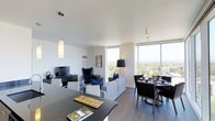 Located at 1101 East Bayaud Avenue in Denver's vibrant Washington Park neighborhood, this single-floor residence offers a sleek, contemporary living experience with an emphasis on natural light and panoramic views. The open-concept layout seamlessly connects the kitchen, living, and dining areas, creating a cohesive and functional flow throughout the space. Large floor-to-ceiling windows dominate the exterior walls, flooding the interior with abundant natural light while framing expansive views of the Denver cityscape and surrounding treetops. The kitchen features a dark, modern countertop that anchors the space and serves as a natural transition into the living area. The living room is styled with a light gray sofa, bold navy accent chairs, and a sleek media console, contributing to a sophisticated yet comfortable ambiance. Adjacent to the living area, the dining space accommodates a round table with dark upholstered chairs, ideal for entertaining. Hardwood-style flooring runs throughout, complementing the crisp white walls and smooth ceilings with recessed and pendant lighting. The overall design aesthetic is clean and modern, with a neutral palette accented by deep navy and black tones.