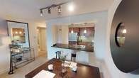 This single-floor residence offers a thoughtfully designed open-concept layout that seamlessly connects the living, dining, and kitchen areas. The kitchen is a standout feature, showcasing rich warm-toned cabinetry, dark granite countertops, a green tile backsplash, and a full suite of stainless steel appliances including a refrigerator, gas range, and built-in microwave. Track lighting illuminates the kitchen, while elegant pendant lights hang above the breakfast bar, which provides additional casual seating and separates the kitchen from the dining area. The dining space accommodates a full-sized table, making it ideal for entertaining. The living room is bright and welcoming, with plush carpeting, neutral walls, and a sliding glass door that opens to a private balcony, allowing natural light to fill the room and offering a pleasant outdoor view. The bedroom spaces are generously sized with soft carpeting, and the primary bedroom features warm accent walls and ample room for full-sized furniture. The bathroom is clean and functional, featuring a vanity with a large mirror, soft lighting, and a tub-shower combination. Throughout the residence, a consistent neutral color palette and thoughtful design choices create a cohesive, comfortable atmosphere.