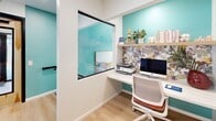Located at 1000 Forest Oak Dr in Charlotte, NC, this single-floor space is a compact yet richly designed interior that makes a strong visual impression. The layout comprises a hallway and a dining room, connected by light hardwood flooring that runs consistently throughout, lending warmth and cohesion to the space.

The hallway is clean and modern, featuring floor-to-ceiling glass partition walls with black framing that offer clear sightlines into an adjacent office area. Natural light filters in from exterior-facing windows, keeping the corridor bright and open. A decorative watercolor landscape painting adds an artistic touch to the passage.

The dining room is the standout feature of the space, distinguished by its vibrant, nature-inspired botanical wallpaper in warm earth and jewel tones that covers an entire accent wall. A built-in banquette upholstered in muted sage green runs along the wall, accented with rust and cream throw pillows. Round wood-topped pedestal tables pair with white wire chairs and upholstered seating to create an inviting, casual dining atmosphere. A statement wire-cage pendant light hangs overhead, adding an industrial-chic touch. A distinctive wood-grain door with black trim anchors one end of the room. Potted greenery throughout adds a fresh, lively energy to this well-curated space.