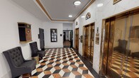 Located at 509 Market St in downtown Shreveport, Louisiana, this distinguished lobby showcases the elegance of a meticulously restored historic building. The space makes an immediate impression with its bold, three-dimensional geometric tile floor rendered in black, white, and terracotta marble — a defining architectural feature that anchors the entire lobby.

Soaring double-height ceilings amplify the grandeur of the main entry area, which is anchored by a dramatic gold lantern chandelier suspended above a central round table topped with a sculptural golden accent piece. Large glass entry doors allow natural light to pour in from Market Street, offering clear views of the surrounding urban streetscape.

White marble wall panels line the elevator corridor, where a bank of polished brass elevator doors adds a refined, Art Deco-inspired touch. Gold crown molding trims the ceiling transitions, reinforcing the building's historic character throughout. A built-in mailbox bank lines one wall of the main corridor, providing functional infrastructure within the elegant setting.

Curated artwork, including large-scale paintings and framed prints, is displayed throughout the lobby, lending a gallery-like quality to the space. Tufted charcoal accent chairs offer seating near the elevator hall, completing this welcoming and architecturally rich entry experience.