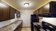 This single-floor unit offers a practical and straightforward layout with distinct living, sleeping, kitchen, and bathroom areas. The galley-style kitchen is efficiently designed with dark espresso cabinetry lining both walls, providing ample upper and lower storage. Laminate countertops in a muted stone finish complement the dark cabinetry, while black appliances — including a refrigerator, electric range with coil burners, and under-cabinet microwave — create a cohesive look. A double stainless steel sink and tile-style flooring complete the kitchen's functional design.

The living area is spacious and open, featuring neutral carpeting and a ceiling fan with integrated lighting. A wall-mounted air conditioning unit provides supplemental cooling, while baseboard heating runs along the perimeter for warmth. The room connects conveniently to the kitchen, creating an open, accessible flow between spaces.

The bedroom offers comfortable proportions with matching neutral carpet, a large window fitted with vertical blinds for natural light and privacy, and baseboard heating. The bathroom includes a full tub-shower combination with a curtain rod, a wood-toned door, and tile flooring. Throughout the unit, warm wood-toned doors and trim add a consistent, cohesive design element.