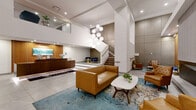 Located on Euclid Avenue in Cleveland, Ohio, this thoughtfully designed two-floor space offers an impressive blend of modern aesthetics and functional amenity areas. The ground floor makes an immediate statement with a grand lobby featuring soaring double-height ceilings, large-format tile flooring, a warm wood-paneled accent wall with a built-in fireplace, and a striking cascading globe chandelier. A staffed reception desk anchors the lobby, complemented by curated mid-century modern seating arrangements atop vibrant area rugs. A dedicated mail and package room with built-in mailboxes and a parcel counter adds everyday convenience. The upper floor transitions to rich hardwood flooring throughout its expansive open layout. Multiple dining areas provide versatile gathering options — one features a long banquette with leather seating, tulip tables, and orange mid-century chairs accented by brass sconce lighting; another offers intimate table seating; and a third is separated by glass partitions for privacy. A lounge area with stylish armchairs and colorful area rugs connects seamlessly to a built-in coffee and refreshment station framed by sleek sliding barn doors. Wide hallways, recessed lighting, and curated artwork throughout both floors contribute to a polished, welcoming atmosphere suited for residents and guests alike.