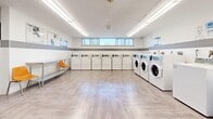 Located at 580 The East Mall in Toronto, Ontario, this shared laundry facility is a thoughtfully designed, single-floor space that offers a clean and organized environment for residents. The room is lined along two walls with a comprehensive array of front-loading and top-loading washing machines alongside matching dryers, providing ample capacity to serve a large number of users efficiently. The machines are numbered and accompanied by clearly posted instructional signage to guide users through the laundry process.

The interior features light wood-effect vinyl flooring laid in a herringbone pattern, contributing to a modern and easy-to-maintain aesthetic. A horizontal band of grey tile runs along the mid-height of the white walls, adding a subtle design element throughout the space. Overhead, long linear fluorescent light fixtures provide bright, even illumination across the entire room.

A row of high, horizontal windows along one wall allows natural light to filter in while maintaining privacy. A stainless steel folding table and orange seating chairs offer a practical waiting and sorting area for users. Additional amenities include a utility sink, a waste bin, and a bulletin board displaying notices and information. A grey entry door provides secure access to the facility. The overall ambiance is clean, bright, and functional, prioritizing practicality and ease of use.