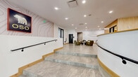 The OSO lobby at 1900 S Acoma St is a generously proportioned, single-floor space that makes a strong architectural statement from the moment you enter. The layout is defined by a seamless blend of polished stone-tile flooring in the entry and hallway areas and warm wood-look flooring throughout the main lounge, creating a clear visual transition between zones.

The central lounge is the heart of the lobby, featuring a large communal dining table surrounded by bold rust-orange upholstered chairs, suspended pendant lighting, and a striking topographic bear mural as a dramatic focal wall. A circular ring chandelier anchors a relaxed seating arrangement with a sage green sofa, armchairs, and an oval coffee table. Curated sculptural artwork, including a vivid red figurative sculpture, adds an artistic energy throughout.

Separate from the main lounge, a quieter seating nook offers olive-green armchairs arranged around a round sculptural table. The mail room is well-appointed with extensive mailbox banks and elegant linear pendant lighting. Restroom facilities are accessed via a mural-adorned corridor. Large windows and glass-paneled doors draw in natural light, while recessed ceiling lighting ensures a consistently bright, welcoming ambiance throughout the entire space.
