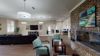 This single-story clubhouse in Mobile, Alabama offers a thoughtfully designed communal space with a welcoming and social atmosphere. The main gathering area serves as the heart of the facility, anchored by a striking floor-to-ceiling brick fireplace flanked by teal upholstered lounge chairs and a rich wood coffee table. Warm hardwood flooring and crown molding run throughout, lending a polished, cohesive look to the interior. A dedicated media lounge features a large wall-mounted flat-screen television and a generously sized sectional leather sofa with reclining seats, complemented by plantation-shuttered windows that allow ample natural light. An adjacent meeting or flex room provides additional seating with a mix of leather recliners and upholstered chairs arranged around a central table, accented by vibrant automotive-themed artwork. A computer nook offers workstations for residents' use. The fully equipped kitchen includes stainless steel appliances, dark cabinetry, granite countertops, a mosaic tile backsplash, and a double sink. The entry area features an exposed brick accent wall and a wooden reception desk, setting a warm and inviting tone throughout the entire clubhouse.
