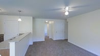 This single-floor residential space offers a straightforward and functional layout designed for comfortable everyday living. The main living area is finished with wall-to-wall carpeting in a neutral tone, providing a warm and inviting atmosphere throughout. A ceiling fan with a built-in light fixture is centrally mounted, offering both air circulation and ambient lighting to the space.

The living area opens naturally toward additional rooms, with two interior doors visible — one leading to a closet or storage area and another providing access to a hallway that connects to further rooms. The hallway, visible in the background, is finished with a smooth ceiling light fixture, guiding the flow through the unit.

The walls throughout are painted in a clean, light neutral tone, lending a bright and airy feel to the interior. White trim and baseboards provide a crisp contrast against the carpeted floors. A partial view of what appears to be a kitchen counter or bar-height surface is visible along one edge of the living area, suggesting a connected or open kitchen layout nearby.

Overall, the space presents a clean, well-maintained interior with a simple and practical design, suitable for a variety of living arrangements.