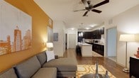 Capital Place at Southwood presents a thoughtfully designed single-floor residence that seamlessly blends functionality with modern style. The open-concept floor plan connects the living area and kitchen, creating a spacious, airy feel that encourages easy movement and interaction throughout the space.

The living area is highlighted by a bold accent wall in warm amber, adding a vibrant splash of color that complements the neutral tones of the room. A ceiling fan overhead provides both comfort and a finishing architectural touch, while the hard surface wood-style flooring runs throughout, lending warmth and visual continuity to the space.

The kitchen is a standout feature, showcasing rich, dark cabinetry that contrasts elegantly with the white countertops and white walls. A central island with bar stool seating makes it ideal for casual dining or entertaining. Pendant lighting above the island adds a refined touch, while black appliances contribute to the kitchen's sleek, cohesive aesthetic.

A hallway from the living area leads to additional rooms, offering a clear and functional layout. The overall ambiance of the residence is modern and inviting, with carefully chosen design elements—warm lighting, contemporary finishes, and a well-coordinated color palette—that come together to create a comfortable, stylish living environment at Capital Place at Southwood.