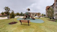 This residential community showcases a blend of classic and contemporary architectural styles. The exterior buildings feature a combination of rich brick facades, stacked stone accents, and warm neutral stucco finishes, with multi-story structures offering private balconies framed by wrought iron railings. Mature trees and carefully maintained landscaping with layered shrubs and ornamental plantings line the building perimeters, creating a lush, welcoming environment.

The outdoor amenity spaces are a central highlight of the community. A resort-style swimming pool serves as the focal point of the courtyard area, surrounded by a spacious brick-paved deck furnished with lounge chairs and shaded dining sets with umbrellas, ideal for relaxation and socializing. Adjacent to the pool, an expansive paver courtyard provides additional outdoor gathering space with seating areas nestled among mature shade trees.

A dedicated children's playground features a multi-level play structure with dual slides, set on a safety-surfaced rubber play area. Park benches nearby provide seating for supervising adults. The surrounding open green lawn areas add to the sense of space and openness throughout the community grounds, creating a comfortable and family-friendly outdoor living environment.