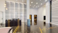 Located at 1401 Spruce Street in Philadelphia's Center City, this grand lobby space presents a seamless blend of classic Art Deco architecture and contemporary design refinements. The entry vestibule is framed by full-height glass panels, creating a luminous transition into the interior while lush greenery flanks the threshold, offering an inviting welcome.

The main lobby floor is clad in richly veined dark marble with gold inlay lettering, anchored by a prominent reception desk in matching black marble with brass accents. Towering fluted columns and floor-to-ceiling sheer drapery panels line the perimeter, diffusing light and lending a sense of grandeur to the expansive open floor plan. Recessed ceiling lighting illuminates the space evenly throughout.

A refined lounge area features a generously scaled sectional sofa in soft blush upholstery, layered with decorative pillows and accented by globe pendant lighting and tropical potted palms, creating a warm and inviting gathering space within the larger lobby.

The elevator banks are finished in warm travertine stone with ornate Art Deco lantern fixtures overhead, polished floors, and brass-toned elevator doors that reflect the building's historic character. The overall ambiance is one of polished sophistication, combining heritage architectural details with modern, curated furnishings.