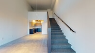 This two-story space showcases a contemporary, loft-inspired design that balances open-plan living with thoughtful functional zones. On the ground floor, polished concrete flooring extends throughout the main living area, which benefits from soaring double-height ceilings and generous natural light admitted through a full-glass entry door and a large sliding door. A high transom window further amplifies the sense of volume and openness.

The kitchen is tucked into a defined alcove and features a warm wood-slat ceiling with track lighting, two-tone cabinetry pairing light wood uppers with deep teal lower cabinets, a subway tile backsplash, dark countertops, and a full suite of stainless steel appliances. A full bathroom on the main level includes a tub-shower combination, a floating vanity, and tile flooring.

A carpeted staircase with a sleek metal handrail leads to the upper level, where an open loft area overlooks the main floor below. This upper space features carpeted flooring, a wall-mounted mini-split system for climate control, and a low half-wall partition at the mezzanine edge. A stacked washer and dryer unit is also present on the upper floor, adding practical utility to the layout.