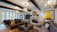 This sophisticated single-floor space presents a thoughtfully designed lounge environment that balances classic architectural details with upscale furnishings. The ceiling draws immediate attention with its bold, dark exposed wood beams contrasting against a bright white surface, while a series of striking gold geometric lantern chandeliers descend at varying heights, casting warm, layered light throughout the open layout.

The primary lounge area centers around rich leather sofas and accent chairs arranged over ornate patterned area rugs atop herringbone-patterned wood-look flooring. A stone-surround fireplace anchors one wall, flanked by a decorative sunburst mirror, while an adjacent dark credenza displays a grid of round gilt-framed mirrors and topiary accents. Wall sconces add additional warmth throughout.

A curved bay window alcove offers an intimate seating vignette with sleek upholstered chairs, a live-edge side table, and floor-to-ceiling windows that invite abundant natural light and views of an outdoor pool and landscaping. Arched corridor openings lead to additional areas, including what appears to be a reception or business center. Tall indoor plants punctuate the space, contributing a fresh, organic element to the overall refined ambiance.