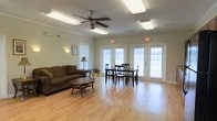This single-floor space offers a bright and functional layout designed for comfort and community. The main gathering area features warm hardwood-style flooring that runs throughout, creating a cohesive and inviting atmosphere. A ceiling fan with an integrated light fixture anchors the room, complementing the recessed panel lighting overhead to ensure the space is well-lit at all times.

The living area is furnished with a plush, dark upholstered sofa and a classic oval coffee table, providing a comfortable seating arrangement ideal for relaxed conversation or lounging. Adjacent to the seating area, a dining set with a dark wood table and matching chairs offers a dedicated space for meals or group activities.

Large windows fitted with white horizontal blinds line the walls, allowing generous amounts of natural light to filter in while maintaining privacy. A pair of French doors with sidelights opens directly to the exterior, enhancing the sense of openness and providing convenient outdoor access. Crown molding adds a refined architectural detail along the ceiling perimeter. The overall ambiance is clean, open, and welcoming, making this a versatile and practical space suited for a variety of communal uses.