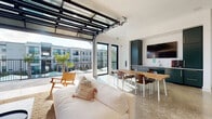 This contemporary single-floor space is defined by its striking architectural centerpiece: a full-width glass-panel overhead door that raises completely to seamlessly merge the interior with a spacious outdoor terrace. When open, the boundary between inside and out dissolves entirely, creating an expansive, light-filled environment with unobstructed views of the surrounding community, palm trees, and a pool area beyond.

The polished concrete floors run throughout, reflecting natural light and reinforcing the clean, modern aesthetic. The living area features a generous white sectional sofa arranged on a woven jute rug, complemented by wood-framed accent chairs and a low-profile coffee table. A large, vibrant abstract artwork anchors one wall, adding a bold splash of color to the otherwise crisp white interior.

The dining area includes a solid wood communal table with seating for several guests, positioned to take full advantage of the open terrace views. Adjacent to the dining space, a built-in kitchenette with dark cabinetry, a wall-mounted television, and a sink provides functional utility. Floor-to-ceiling black-framed glass doors and windows line the perimeter, ensuring abundant natural light even when the overhead door is closed. The overall ambiance is airy, open, and modern — well-suited for both social gatherings and relaxed lounging.