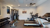 This dedicated fitness space is thoughtfully designed to support a well-rounded exercise routine. The room features a clean, functional layout with durable carpet flooring suited for high-activity use. Cardio equipment takes center stage, with a treadmill and an elliptical trainer positioned to allow ample room for movement. A multi-station cable strength training machine is situated along one wall, providing a comprehensive range of resistance exercise options alongside a flat workout bench. The ceiling is fitted with recessed lighting that ensures bright, even illumination throughout the space, while a ceiling fan promotes airflow and comfort during intense workouts. A subtle decorative touch — a framed landscape print mounted on the white wall — adds a calm, motivating element to the environment. The overall design is straightforward and practical, with white walls and a neutral color palette that keep the focus on functionality. The space is self-contained on a single floor, making it easily accessible and convenient for regular use.