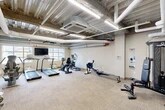 Located at 16 Cinema Dr in Lansing, NY, this fitness room offers a comprehensive exercise facility within a compact, efficiently utilized single-floor layout. The space features an industrial aesthetic defined by an exposed concrete ceiling with visible ductwork, large white pipes, and linear LED lighting that illuminates the room evenly throughout.

The exercise room is outfitted with a diverse selection of cardio equipment, including multiple treadmills, an elliptical trainer, a recumbent stationary bike, and Concept2 rowing machines, providing users with a full range of aerobic workout options. A dedicated strength training area includes a multi-functional cable machine, a flat workout bench, and a dumbbell rack stocked with free weights to support resistance training needs.

Natural light enters the space through several windows, adding a bright and open feel to the room. A wall-mounted flat-screen television is positioned near the cardio equipment for entertainment during workouts. Additional amenities include a bulletin board, a hand sanitizer dispenser, a water cooler, and a recycling bin, reflecting a thoughtful approach to user convenience. The floor is covered with durable, neutral-toned carpet tiles suited for a high-traffic fitness environment. The overall ambiance is functional and practical, designed to support a variety of fitness routines.