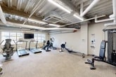 Located at 16 Cinema Dr in Lansing, NY, this fitness room offers a comprehensive exercise facility within a compact, efficiently utilized single-floor layout. The space features an industrial aesthetic defined by an exposed concrete ceiling with visible ductwork, large white pipes, and linear LED lighting that illuminates the room evenly throughout.

The exercise room is outfitted with a diverse selection of cardio equipment, including multiple treadmills, an elliptical trainer, a recumbent stationary bike, and Concept2 rowing machines, providing users with a full range of aerobic workout options. A dedicated strength training area includes a multi-functional cable machine, a flat workout bench, and a dumbbell rack stocked with free weights to support resistance training needs.

Natural light enters the space through several windows, adding a bright and open feel to the room. A wall-mounted flat-screen television is positioned near the cardio equipment for entertainment during workouts. Additional amenities include a bulletin board, a hand sanitizer dispenser, a water cooler, and a recycling bin, reflecting a thoughtful approach to user convenience. The floor is covered with durable, neutral-toned carpet tiles suited for a high-traffic fitness environment. The overall ambiance is functional and practical, designed to support a variety of fitness routines.