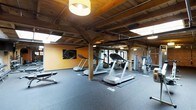 This fitness center combines industrial-rustic character with modern functionality. The space is defined by its striking exposed wood beam ceiling supported by natural timber columns, which lend a warm, lodge-like aesthetic that sets it apart from a typical gym environment. Complementing the woodwork, exposed brick walls add texture and depth to the space, while bold amber-toned accent walls contribute a vibrant, energizing atmosphere.

The flooring consists of durable rubber gym matting, providing both safety and comfort throughout the workout area. The space is generously sized, offering ample room between equipment to move freely. Cardio equipment is prominently featured, including multiple treadmills and stationary bikes, arranged to allow users a sense of openness during their workout. Strength and resistance training machines are also available, positioned along the perimeter to maximize floor space efficiency.

Lighting is provided by a combination of pendant fixtures and ambient overhead sources, ensuring the space is well-lit without feeling harsh. Large windows allow natural light to filter into the room, enhancing the overall brightness and creating an inviting environment. A wall of mirrors expands the visual space and supports users during exercise. Overall, the fitness center delivers a functional, well-appointed workout experience within a characterful and thoughtfully designed setting.