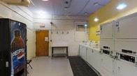 This single-floor shared laundry facility is designed with functionality and practicality in mind. The room features a row of white top-load washing machines and stacked dryer units lining the right side, providing ample laundry capacity for multiple users. A centrally placed utility folding table offers a convenient surface for sorting and folding clothes. The space is brightened by a bold yellow accent wall on one end, adding a touch of color to an otherwise utilitarian environment. Overhead, a ceiling fan aids in air circulation, while pendant lighting and recessed fixtures ensure the room is well-lit. The floor is finished with a durable gray surface, complemented by a rubber anti-fatigue mat in front of the machines. An exposed pipe system runs along the ceiling, consistent with the practical, no-frills aesthetic of the space. A wall-mounted air conditioning unit provides climate control, and a standard entry door with a posted notice offers secure access. Hooks along one wall provide additional storage convenience. An illuminated exit sign ensures clear safety compliance throughout the room.