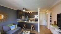 This single-floor unit showcases an open-concept layout that seamlessly connects the kitchen and living area, creating a cohesive and functional living space. The kitchen is a standout feature, outfitted with rich dark espresso cabinetry that extends to the upper and lower sections, providing ample storage. Stainless steel appliances, including a refrigerator, range, and built-in microwave, complement the modern aesthetic. The kitchen's granite countertops feature warm beige and grey tones, adding texture and elegance to the space.

A peninsula counter with bar seating accommodates two wooden stools, serving as a natural divider between the kitchen and living area while offering a casual dining option. A mounted wine glass rack adds a functional yet stylish touch to the kitchen wall. Crown moulding details along the ceiling add a refined architectural element throughout.

The living area features warm hardwood-style flooring that runs continuously, unifying the space. A flush-mount ceiling light fixture and track lighting in the kitchen ensure well-distributed illumination. A hallway visible in the background leads to additional areas of the unit, hinting at further functional space beyond the main open-plan area.