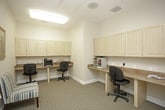 This single-floor business center is thoughtfully designed to provide a functional and productive workspace. The room features multiple built-in workstations with stone-topped counters and light wood cabinetry overhead, offering ample desk space and storage for office essentials. Two ergonomic task chairs are positioned at a shared workstation equipped with a multifunction printer, making it well-suited for printing, copying, and scanning tasks. A dedicated mail and document sorting station with a multi-slot organizer sits along one wall, keeping correspondence and paperwork neatly arranged. A separate workstation tucked into a corner alcove provides a more private setting, complete with its own desk, task chair, and a cork bulletin board for notes and reminders. The space is finished with neutral-toned walls, textured wall-to-wall carpeting, and bright overhead lighting, including a skylight that enhances the overall brightness of the room. A comfortable striped accent chair adds a welcoming touch, and a framed landscape painting contributes a warm, polished aesthetic. Glass-paneled doors connect the business center to an adjoining corridor, allowing for easy accessibility while maintaining a sense of enclosure and focus within the workspace.