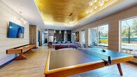 Located at 155 Ames St in Marlborough, MA, this thoughtfully designed amenity space offers a dynamic and upscale environment for recreation and socializing. The main game room is the centerpiece, showcasing wood-plank flooring, a striking tray ceiling finished in warm golden wood tones, and a cluster of modern globe pendant lights that lend a sophisticated ambiance. The space is well-appointed with a full-size pool table, ping-pong table, and a shuffleboard table, catering to a variety of leisure activities. Large sliding glass doors and floor-to-ceiling windows flood the room with natural light and provide direct views of an outdoor patio area. A stunning black marble-topped bar island with fluted wood paneling and teal velvet barstools anchors the kitchen area, which features dark cabinetry with gold hexagonal tile accents and stainless steel appliances. A separate indoor golf simulator room offers a high-definition projection screen, artificial turf mat, and large windows overlooking lush greenery. Cozy lounge nooks with plush blue velvet sectional sofas, warm wood accent walls, and curated sculptural artwork provide intimate retreat spaces throughout. The overall atmosphere is modern, vibrant, and resort-inspired.