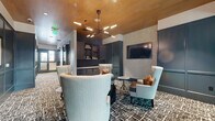 This single-floor amenity space in Colorado Springs, Colorado, is designed with a refined, contemporary aesthetic that balances comfort and functionality. The main lounge area serves as the centerpiece, featuring an arrangement of wingback chairs in soft blue-gray and warm neutral tones, paired with marble-topped accent tables and a bold dark coffee table. A striking sputnik-style chandelier hangs from a warm wood-paneled ceiling, casting ambient light across the patterned, multi-tonal carpet that grounds the entire space.

One wall is finished in deep teal paneling, adorned with a curated set of framed artwork, while textured gray wallcovering on adjacent walls adds visual depth. Built-in dark cabinetry with open shelving and a flat-panel television mounted to the wall provide both storage and entertainment functionality. Glass-paneled partitions divide the main lounge from a semi-private conference area, which includes a round marble table, cognac leather chairs, and a wall-mounted display screen. A dedicated individual workstation tucked into a teal-paneled alcove features pendant lighting and a built-in white desk surface. Together, these areas create a polished, versatile environment suited for both collaborative gatherings and focused individual work.