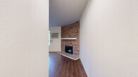 Located at 6104 Cahuenga Blvd in the North Hollywood neighborhood of Los Angeles, this single-floor space offers a well-organized layout centered around a bright, open living area. The living room serves as the focal point of the space, featuring a striking full-height exposed brick fireplace with a white mantel that adds a classic, rustic charm to the interior. Rich, dark hardwood-style flooring extends throughout the space, providing warmth and visual continuity between areas. A row of large windows along one wall floods the living area with abundant natural light and offers views of the surrounding neighborhood. A wall-mounted air conditioning unit ensures year-round comfort. The open layout connects the living area to an adjacent kitchen, which includes white cabinetry and countertops, offering functional utility in a compact footprint. A secondary room, equipped with a ceiling fan and light fixture, provides additional flexible living space. Clean white walls throughout create a neutral, versatile backdrop. The overall ambiance is bright, functional, and characterized by the interplay of classic architectural details—most notably the prominent brick fireplace—with practical, modern finishes.