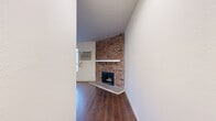 Located at 6104 Cahuenga Blvd in the North Hollywood neighborhood of Los Angeles, this single-floor space offers a well-organized layout centered around a bright, open living area. The living room serves as the focal point of the space, featuring a striking full-height exposed brick fireplace with a white mantel that adds a classic, rustic charm to the interior. Rich, dark hardwood-style flooring extends throughout the space, providing warmth and visual continuity between areas. A row of large windows along one wall floods the living area with abundant natural light and offers views of the surrounding neighborhood. A wall-mounted air conditioning unit ensures year-round comfort. The open layout connects the living area to an adjacent kitchen, which includes white cabinetry and countertops, offering functional utility in a compact footprint. A secondary room, equipped with a ceiling fan and light fixture, provides additional flexible living space. Clean white walls throughout create a neutral, versatile backdrop. The overall ambiance is bright, functional, and characterized by the interplay of classic architectural details—most notably the prominent brick fireplace—with practical, modern finishes.