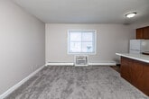Located at 2013 W First St in Ankeny, Iowa, this one-bedroom, one-bathroom unit offers a practical and efficiently arranged single-floor layout. The open living and kitchen area is anchored by a peninsula counter with a white laminate top, providing a natural division between the carpeted living space and the kitchen. The kitchen features warm wood cabinetry — both upper and lower — along with a white refrigerator, range, and a double sink, all set on tile flooring. A window with blinds allows natural light to fill the living area, which is also equipped with a wall-mounted air conditioning unit and baseboard heating. The bedroom is a comfortable, carpeted room with its own window overlooking the exterior and a built-in closet with double doors and a double hanging rod, offering useful storage. The bathroom includes a full-size tub and shower surround, a toilet, and a vanity sink. Neutral gray walls and carpet tones are consistent throughout, giving the unit a cohesive and understated feel. Baseboard heaters are present in multiple rooms, supporting efficient temperature control. This unit is a straightforward, functional space well-suited for a single occupant or couple seeking a convenient location in Ankeny.