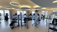This contemporary fitness center offers a thoughtfully designed workout environment across a single floor. The space is outfitted with a comprehensive selection of professional-grade exercise equipment, including treadmills, stationary bikes, a recumbent bike, and a variety of strength training machines — providing everything needed for a full-body workout routine.

The interior design is sleek and modern, with a neutral color palette of soft whites and warm beige tones that create a clean, energizing atmosphere. The ceiling is a standout architectural feature, adorned with large, circular recessed panels paired with elegant ring-shaped pendant lights that cast a warm, ambient glow throughout the space, enhancing both aesthetics and functionality.

Large floor-to-ceiling windows line the perimeter of the room, flooding the interior with natural light and offering expansive views of the surrounding landscape. The smooth, light-toned flooring complements the overall design while providing a practical, easy-to-maintain surface suited for high-traffic use.

Overall, the fitness center strikes a balance between form and function — delivering a polished, upscale environment that is both visually impressive and highly practical for users seeking a premium workout experience.