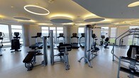 This contemporary fitness center offers a thoughtfully designed workout environment across a single floor. The space is outfitted with a comprehensive selection of professional-grade exercise equipment, including treadmills, stationary bikes, a recumbent bike, and a variety of strength training machines — providing everything needed for a full-body workout routine.

The interior design is sleek and modern, with a neutral color palette of soft whites and warm beige tones that create a clean, energizing atmosphere. The ceiling is a standout architectural feature, adorned with large, circular recessed panels paired with elegant ring-shaped pendant lights that cast a warm, ambient glow throughout the space, enhancing both aesthetics and functionality.

Large floor-to-ceiling windows line the perimeter of the room, flooding the interior with natural light and offering expansive views of the surrounding landscape. The smooth, light-toned flooring complements the overall design while providing a practical, easy-to-maintain surface suited for high-traffic use.

Overall, the fitness center strikes a balance between form and function — delivering a polished, upscale environment that is both visually impressive and highly practical for users seeking a premium workout experience.