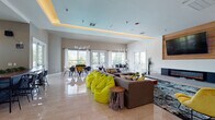 This dynamic single-floor community space is designed to inspire connection and comfort through a thoughtfully open-concept layout. The main lounge area is anchored by a sleek wall-mounted fireplace set against a warm wood panel, with a large flat-screen TV above. Plush sectional seating, vibrant yellow-green bean bag chairs, and colorful accent furniture create an inviting, energetic atmosphere throughout. A striking living green wall bearing the "YE" branded logo serves as a bold focal point, adding a natural element to the contemporary interior. Polished large-format tile flooring runs throughout the space, reflecting abundant natural light streaming in through floor-to-ceiling glass doors and generous windows. The dining and gaming area is outfitted with stylish pendant lighting and direct access to an outdoor terrace, where additional seating extends the living experience into the open air. A separate coworking zone features long communal tables, mixed seating, and a mounted display screen, ideal for collaborative work or study. Tray ceilings with warm cove lighting lend architectural elegance across multiple zones. A kitchenette area with cabinetry provides convenient utility. The overall ambiance balances modern sophistication with playful, welcoming energy throughout every corner of this well-appointed space.