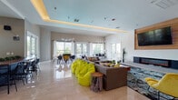This dynamic single-floor community space is designed to inspire connection and comfort through a thoughtfully open-concept layout. The main lounge area is anchored by a sleek wall-mounted fireplace set against a warm wood panel, with a large flat-screen TV above. Plush sectional seating, vibrant yellow-green bean bag chairs, and colorful accent furniture create an inviting, energetic atmosphere throughout. A striking living green wall bearing the "YE" branded logo serves as a bold focal point, adding a natural element to the contemporary interior. Polished large-format tile flooring runs throughout the space, reflecting abundant natural light streaming in through floor-to-ceiling glass doors and generous windows. The dining and gaming area is outfitted with stylish pendant lighting and direct access to an outdoor terrace, where additional seating extends the living experience into the open air. A separate coworking zone features long communal tables, mixed seating, and a mounted display screen, ideal for collaborative work or study. Tray ceilings with warm cove lighting lend architectural elegance across multiple zones. A kitchenette area with cabinetry provides convenient utility. The overall ambiance balances modern sophistication with playful, welcoming energy throughout every corner of this well-appointed space.