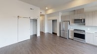 This single-floor unit showcases a thoughtfully designed open layout with a cohesive modern aesthetic carried throughout. Wood-look plank flooring runs continuously across the living areas, creating a warm yet contemporary foundation. The open-concept living and kitchen area is bright and airy, with elevated ceilings and recessed lighting adding to the sense of spaciousness.

The kitchen is well-appointed with a full suite of stainless steel appliances, including a French door refrigerator, gas range, over-the-range microwave, and dishwasher. Light wood flat-front cabinetry extends to ceiling height, maximizing storage, while a subway tile backsplash and light-toned countertops complete the refined look. A built-in sink sits neatly beneath upper cabinets, maintaining the kitchen's clean lines.

The living area benefits from natural light through multiple windows offering urban views, complemented by a ceiling fan and recessed lighting. A sliding barn door provides stylish access to closet space, while additional built-in closets offer generous storage throughout.

The bathroom features light wood vanity cabinetry, a white quartz countertop, and a large mirror, maintaining the unit's neutral, modern palette. The space as a whole presents a clean, move-in ready environment with consistent finishes and functional design throughout.
