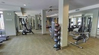 This fitness center offers a comprehensive workout environment spread across a single floor. The space is anchored by carpeted flooring and supported by structural columns that divide the room into distinct workout zones without compromising the open feel. Ceiling fans are mounted overhead, promoting air circulation throughout the facility, while recessed lighting ensures consistent illumination across all areas.

The cardio section features stationary bikes and elliptical machines, positioned to allow comfortable use. A medicine ball rack holds a full range of weighted balls, catering to functional and core training. Additional strength and resistance machines are distributed throughout the space, providing options for a full-body workout routine.

Large mirrors are mounted along the walls, visually expanding the room and allowing users to monitor their form during exercise. A wall-mounted television is also present, offering entertainment during workouts. Natural light filters in through windows visible toward the perimeter, adding brightness to the interior. The overall ambiance is clean, functional, and well-organized, making it a practical and inviting space for fitness activities.