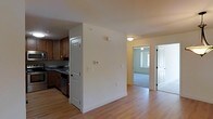 This single-floor residential space offers a well-organized and functional layout designed for comfortable everyday living. The kitchen is outfitted with warm wood cabinetry, dark countertops, and stainless steel appliances, providing ample storage and workspace for meal preparation. A partial wall separates the kitchen from the adjacent living area, creating a defined yet open feel throughout the main living space.

Light hardwood flooring extends across the main areas, adding warmth and continuity to the interior. The neutral white and cream wall tones keep the space feeling bright and airy. A hallway connects the main living area to additional rooms, including what appears to be a carpeted bedroom with a window that allows natural light to filter in, contributing to an inviting atmosphere.

Multiple doorways are visible throughout, suggesting a layout that balances privacy and accessibility between rooms. The overall design is straightforward and practical, with clean lines and minimal ornamentation. The unit benefits from ceiling-mounted light fixtures that provide consistent illumination throughout. This space is well-suited for those seeking a tidy, move-in-ready home with a logical floor plan and functional amenities across its single level.